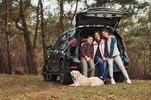 família feliz se diverte com seu cachorro perto de carro moderno ao ar livre na floresta foto