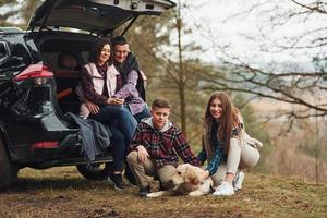 família feliz sentada e se divertindo com seu cachorro perto de carro moderno ao ar livre na floresta foto