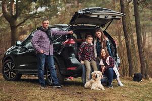 família feliz se diverte com seu cachorro perto de carro moderno ao ar livre na floresta foto