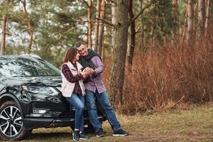 casal maduro positivo parado perto de seu carro com bebida quente nas mãos. ao ar livre na floresta foto