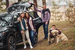 família feliz se diverte com seu cachorro perto de carro moderno ao ar livre na floresta foto
