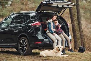 menina alegre com seu irmão senta-se na parte de trás do carro moderno com seu cachorro ao ar livre na floresta foto