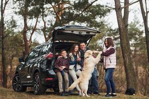 família feliz se diverte com seu cachorro perto de carro moderno ao ar livre na floresta foto