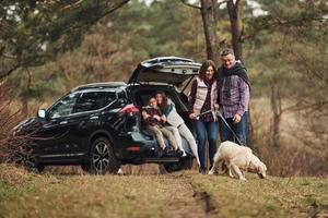 família feliz se diverte com seu cachorro perto de carro moderno ao ar livre na floresta foto