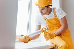 faz-tudo de uniforme amarelo trabalha com cola para janela dentro de casa. concepção de renovação de casa foto