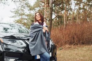 mulher em pé perto de carro preto moderno ao ar livre na floresta foto