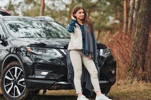 jovem alegre em pé perto de carro preto moderno ao ar livre na floresta foto