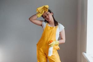 dona de casa cansada em pé de uniforme amarelo com spray de limpeza para janelas dentro de casa. concepção de renovação de casa foto