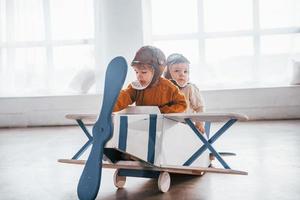 dois meninos em uniforme de piloto retrô se divertindo com avião de brinquedo dentro de casa foto