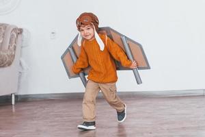 menino em uniforme de piloto retrô correndo com avião de brinquedo dentro de casa foto