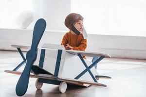 garotinho em uniforme de piloto retrô se divertindo com avião de brinquedo dentro de casa foto