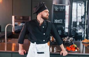 cozinheiro profissional jovem chef de uniforme em pé perto da mesa e posando para uma câmera na cozinha foto