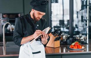 cozinheiro profissional jovem chef de uniforme em pé com o bloco de notas na cozinha foto