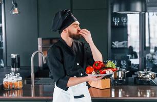 cozinheiro profissional jovem chef de uniforme segura prato com legumes na cozinha foto