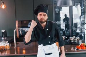 cozinheiro profissional jovem chef de uniforme em pé perto da mesa e posando para uma câmera na cozinha foto