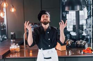 cozinheiro profissional jovem chef de uniforme em pé perto da mesa e posando para uma câmera na cozinha foto