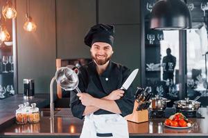 cozinheiro profissional jovem chef de uniforme em pé perto da mesa e posando para uma câmera na cozinha foto