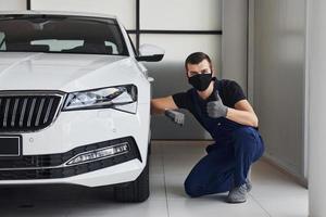 cara de uniforme azul e máscara protetora preta sentado perto de um carro branco fixo e mostra o polegar para cima foto