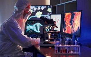 monitores com informações sobre a mesa. cientista em uniforme protetor branco trabalha com coronavírus e tubos de sangue em laboratório foto