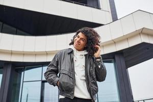 jovem bonito com cabelo preto encaracolado posando para a câmera na rua contra a construção foto