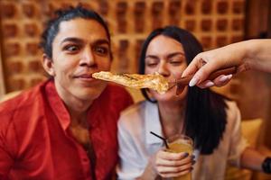casal jovem multiétnico sentado dentro de casa juntos e comendo pizza foto