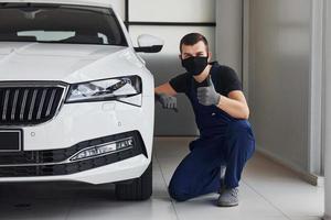 cara de uniforme azul e máscara protetora preta sentado perto de um carro branco fixo e mostra o polegar para cima foto