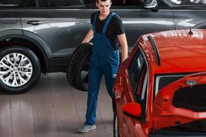 trabalhador de uniforme preto e azul segura a roda do carro e caminha com um automóvel quase vermelho foto