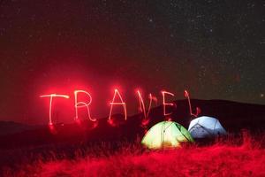 palavra de viagem de néon vermelho. duas tendas iluminadas sob as estrelas nas montanhas à noite foto