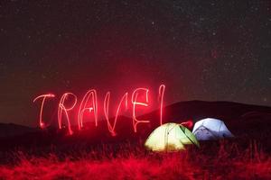 palavra de viagem de néon vermelho. duas tendas iluminadas sob as estrelas nas montanhas à noite foto
