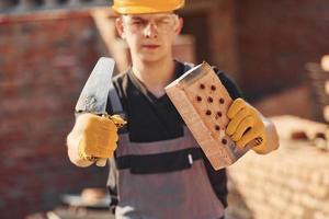 retrato de trabalhador da construção civil em uniforme e equipamento de segurança que está de pé na construção e segurando tijolo e ferramenta foto