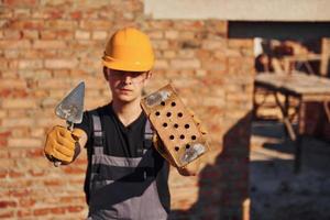 retrato de trabalhador da construção civil em uniforme e equipamento de segurança que está de pé na construção e segurando tijolo e ferramenta foto
