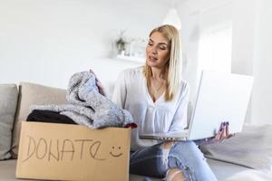 mão de mulher segurando a caixa de doação com roupas e laptop. jovem mulher com caixa de doação em casa. caixa de doação para pobres com roupas em mãos femininas. foto