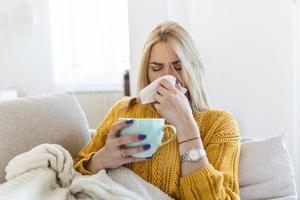 mulher.flu.mulher doente pegou resfriado. espirrar no tecido. dor de cabeça. vírus .medicina. jovem infectada com frio assoando o nariz no lenço. mulher doente com dor de cabeça sentada em um sofá foto