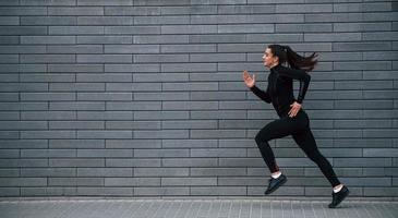 jovem esportiva em roupas esportivas pretas correndo ao ar livre perto da parede cinza foto