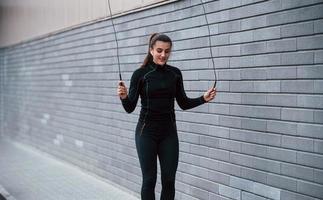 jovem esportiva em roupas esportivas pretas praticando com pular corda ao ar livre perto da parede cinza foto