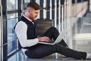 jovem passageiro do sexo masculino em elegantes roupas formais, sentado com o laptop no saguão do aeroporto foto