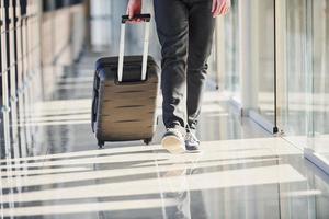 visão aterrada do passageiro do sexo masculino em elegantes roupas formais está no saguão do aeroporto andando com bagagem foto