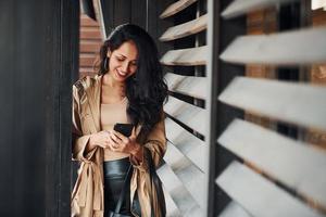 mulher com cabelo preto encaracolado em pé perto de janelas de madeira com o telefone nas mãos foto