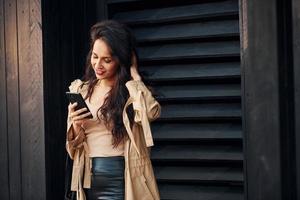 mulher com cabelo preto encaracolado em pé contra o exterior do edifício de madeira preta e usando o telefone foto