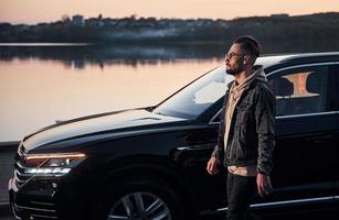 homem bonito sem barba em roupas da moda caminha perto de seu carro preto à noite foto