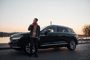 bonito homem sem barba em roupas da moda parado perto de seu carro preto e fumando foto