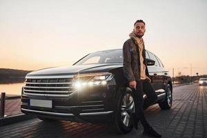 bonito homem sem barba em roupas da moda parado perto de seu carro preto e fumando foto