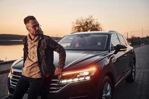 homem bonito com a barba por fazer em roupas da moda, apoiando-se em seu carro preto foto