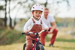 pai de camisa branca ensinando a filha a andar de bicicleta ao ar livre foto