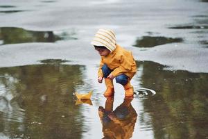 garoto com manto impermeável amarelo e botas brincando com brinquedo de barco de papel feito à mão ao ar livre depois da chuva foto