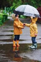 duas crianças com guarda-chuva em mantos impermeáveis amarelos e botas brincando ao ar livre depois da chuva juntos foto