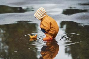 garoto com manto impermeável amarelo e botas brincando com brinquedo de barco de papel feito à mão ao ar livre depois da chuva foto