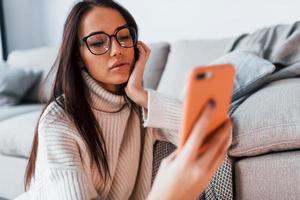 jovem mulher bonita em óculos sentado em casa sozinho com o telefone nas mãos foto