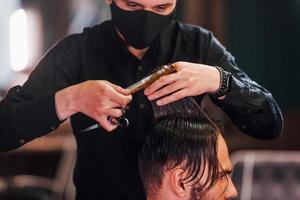 jovem barbudo sentado e cortando o cabelo na barbearia por cara com máscara protetora preta foto