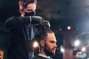 jovem barbudo sentado e cortando o cabelo na barbearia por cara com máscara protetora preta foto